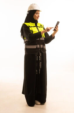دمج التكنولوجيا الحديثة بالهندسة، العمل في القطاع الهندسي, بورتريه من الجانب لمهندسة عربية خليجية سعودية ترتدي خوذة وسترة الحماية تقوم بمتابعة سير الاعمال عن بعد، شابة سعودية تحمل بيدها الهاتف المحمول، خلفية بيضاء