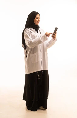 تصفح الانترنت عبر الهاتف، بورتريه لطبيبة عربية خليجية سعودية ترتدي المعطف الأبيض والسماعة تحمل بيدها الجوال، تقديم الخدمة والاستشارة الطبية، مهن ووظائف نسائية،استخدام التكنولوجيا الحديثة، الجسم بالطول الكامل، خلفية بيضاء