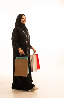 Enjoying shopping and getting discounts, a side portrait of a Saudi Arabian Gulf woman wearing a black abaya, holding paper bags in her hands, looking with gestures of joy and happiness, a full-body image, white background.