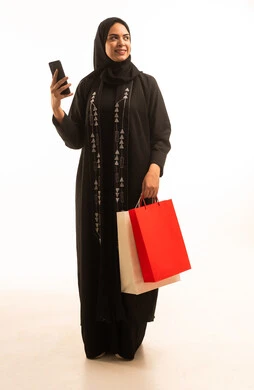 A portrait of a Saudi Arabian Gulf woman wearing a black abaya and a hijab, holding colorful shopping bags in her hands, looking at something, taking advantage of seasonal offers and discounts, with expressions and gestures of joy, full-body shot, white background.