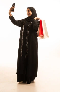 اجراء مكالمة فيديو هاتفية باستخدام الهاتف المحمول، بورتريه لإمرأة عربية خليجية سعودية محجبة ترتدي عباءة سوداء تحمل بيديها أكياس التسوق الملونة، الاستفادة من العروض والتخفيضات الموسميه، تعابير وايماءات السرور، الجسم بالطول الكامل، خلفية بيضاء