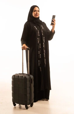 Looking at the camera with gestures of joy, a side portrait of a Saudi Gulf Arab woman wearing a black abaya holding a travel bag and a mobile phone, full-body image, white background.