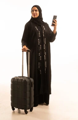 Looking at something, a portrait of a Saudi Arabian Gulf woman wearing a black abaya holding a travel bag and a mobile phone, hotel booking through a travel app, chatting on social media, full-body image, white background.