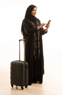 الحجز الفندقي عبر تطبيق السفر, بورتريه لإمرأة عربية خليجية سعودية ترتدي العباءة السوداء تمسك حقيبة السفر والهاتف المحمول بإيماءات السرور، الدردشة عبر وسائل التواصل الاجتماعي, صورة الجسم بالطول الكامل, خلفية بيضاء