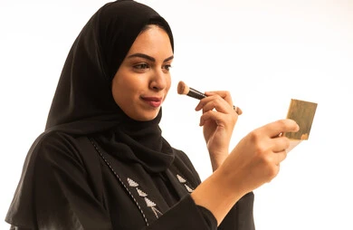 The interest in beauty and appearance, a close-up portrait of a Saudi Arabian Gulf woman wearing a black abaya, using a blush brush, enjoying putting on makeup, the concept of adornment and beautification, white background.