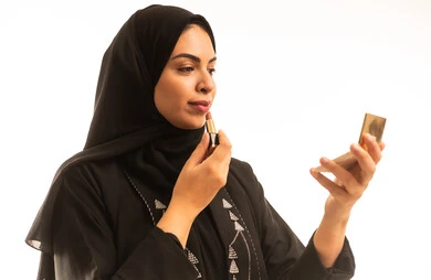 A close-up portrait of a Saudi Arabian Gulf woman wearing a black abaya and applying lipstick, enjoying putting on makeup, caring about beauty and appearance, the concept of adornment and beautification, with a white background.