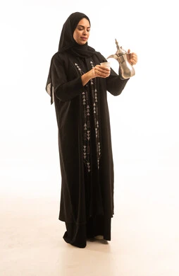 The concept of generosity and hospitality, a portrait of a young Arab Gulf Saudi woman wearing a black abaya pouring coffee from a brass coffee pot, on the day we began in 1727 AD, the anniversary of the founding of the first Saudi state on February 22, with a white background.