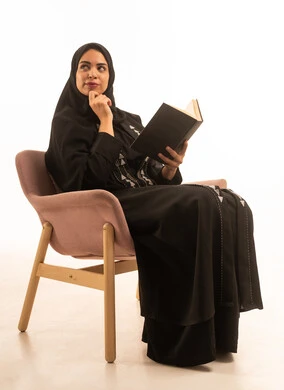 Looking at a place, the culture of self-learning, spending time learning and educating oneself, enjoying reading books, a portrait of a Saudi Arabian Gulf woman wearing a black abaya, sitting on a chair holding a book in her hands, with a white background.