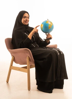 A portrait of a Saudi Arabian Gulf woman wearing a black abaya sitting on a comfortable chair pointing to a globe, exploring and researching around the world, the concept of elegance and taking care of one's appearance, with a white background.