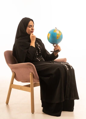 Confusion and thinking about something, embarking on an exploratory tourist trip around the world, a portrait of a Gulf Arab Saudi woman wearing a black abaya sitting on a comfortable chair looking at a globe, white background.