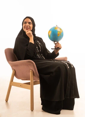 البحث والإستكشاف حول العالم، بورتريه لامرأة عربية خليجية سعودية ترتدي العباءة السوداء تجلس على كرسي مريح تحمل مجسم الكرة الارضية تنظر الى شىء ما, مفهوم الأناقة والاعتناء بالمظهر الخارجي، خلفية بيضاء