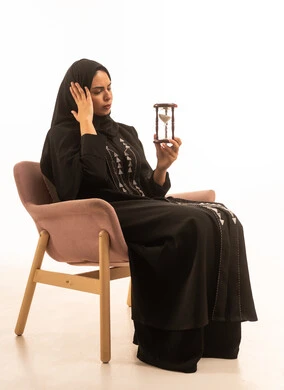 التخطيط لتحقيق الأهداف في المستقبل، القيام بمراقبة توقيت الرمل, إدراك قيمة اللحظات الثمينة, بورتريه لامرأة عربية خليجية سعودية محجبة ترتدي عباءة سوداء تجلس على كرسي مريح و تمسك ساعة رملية وتضع يدها على خدها, التحليل والتدقيق واتخاذ القرارات, خلفية بيضاء