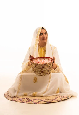 A portrait of a young Arab Gulf Saudi woman wearing a white abaya sitting on a straw mat in front of a basket full of flowers, looking at the camera with gestures of joy, commemorating the founding of the first Saudi state on February 22, local product and harvest, white background.