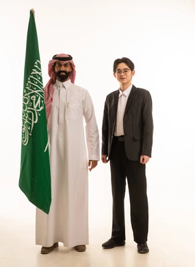 بورتريه لرجل عربي خليجي سعودى يرتدي الثوب التقليدى والشماغ يحمل علم المملكة بجانب رجل اسيوى يرتدي البدلة الرسمية ينظران الى الكاميرا بفخرو ثقة، تبادل الثقافة بين البلاد, إحياء ذكرى اليوم الوطني السعودي 23 سبتمبر، خلفية بيضاء