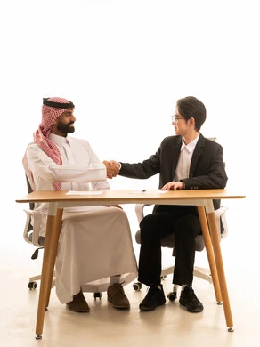 A portrait of a Saudi Gulf Arab man wearing traditional attire and a shemagh sitting at a desk in a business meeting, shaking hands with a young Asian man in a formal suit, gestures indicating happiness at the success of the deal, networking and building social relationships, white background.