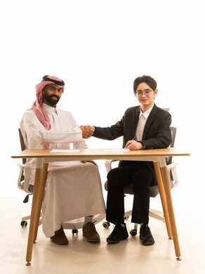 Gestures indicating happiness at the success of the deal, a portrait of a Saudi Gulf Arab man wearing traditional attire and a ghutrah sitting at a desk in a business meeting with a young Asian man in a formal suit, shaking hands, getting to know each other, and building social relationships, white background.