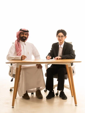 تقديم الأفكار وشرح تفاصيل وثغرات المشروع، بورتريه لرجل عربى خليجى سعودى يرتدى الثوب التقليدى والشماغ يجلس على مكتب في اجتماع عمل مع شاب اسيوى يرتدي البدلة الرسمية يتناقشان في مخطط الشركة، خلفية بيضاء