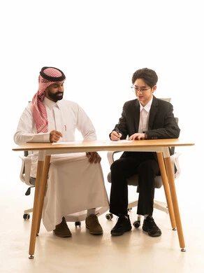 بورتريه لرجل عربى خليجى سعودى يرتدى الثوب التقليدى والشماغ يجلس على مكتب في اجتماع عمل مع شاب اسيوى يرتدي البدلة الرسمية يتناقشان في مخطط الشركة،  تقديم الأفكار وشرح تفاصيل وثغرات المشروع، خلفية بيضاء