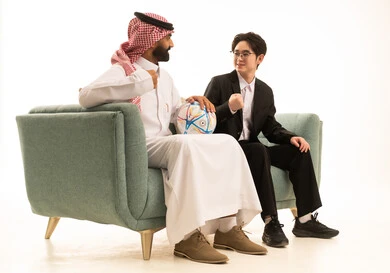 الفرح لفوز الفريق المفضل، بورتريه من الجانب لرجل عربى خليجى سعودى يرتدى الثوب التقليدى والشماغ يجلس على الاريكة بجانب شاب اسيوى يرتدى البدلة الرسمية بجانبهم كورة قدم، خلفية بيضاء