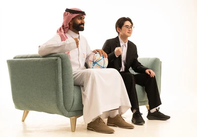 قضاء وقت ممتع بمتابعة المباراة, رفع قبضة اليد بتعابير الفوز والانتصار، بورتريه من الجانب لرجل عربى خليجى سعودى يرتدى الثوب التقليدى والشماغ يجلس على الاريكة بجانب شاب اسيوى يرتدى البدلة الرسمية بجانبهم كورة قدم، خلفية بيضاء