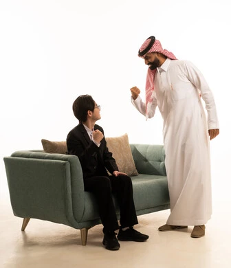 بورتريه من الجانب لشاب اسيوى يرتدى البدلة الرسمية يجلس على الاريكة امام رجل عربى خليجى سعودى يرتدى الثوب التقليدى والشماغ يقف بحماس، مفهوم الاندماج بمشاهدة الفريق المفضل، رفع قبضة اليد بتعابير الفوز والانتصار، قضاء اوقات الفراغ الممتعة ، خلفية بيضاء