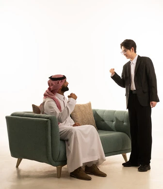 مفهوم الاندماج بمشاهدة الفريق المفضل، رفع قبضة اليد بتعابير الفوز والانتصار، قضاء اوقات الفراغ الممتعة ، بورتريه من الجانب لرجل عربى خليجى سعودى يرتدى الثوب التقليدى والشماغ يجلس على الاريكة بجانب شاب اسيوى يرتدى البدلة الرسمية يقف بحماس، خلفية بيضاء