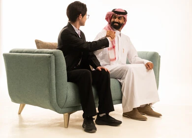 تشجيع فريق كورة القدم المفضل، رفع قبضة اليد بتعابير الفوز والانتصار، بورتريه من الجانب لرجل عربى خليجى سعودى يرتدى الثوب التقليدى والشماغ يجلس على الاريكة بجانب شاب اسيوى يرتدى البدلة الرسمية، خلفية بيضاء