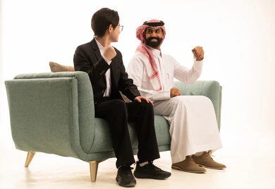 رفع اليد بتعابير الفوز، بورتريه من الجانب لرجل عربى خليجى سعودى يرتدى الثوب التقليدى والشماغ يجلس على الاريكة بجانب شاب اسيوى يرتدى البدلة الرسمية يبتسمان، خلفية بيضاء