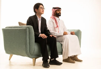 القيام بمشاهدة شىء ما، بورتريه من الجانب لرجل عربى خليجى سعودى يرتدى الثوب التقليدى والشماغ يجلس على الاريكة بجانب شاب اسيوى يرتدى البدلة الرسمية ينظران الى المباراه بتعابير الاندماج، خلفية بيضاء