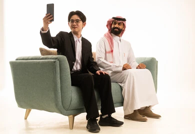 التقاط السيلفى عبر الهاتف المحمول، بورتريه لرجل عربى خليجى سعودى يرتدى الثوب التقليدى والشماغ يجلس على الاريكة بجانب شاب اسيوى يرتدى البدلة الرسمية يمسك الجوال، خلفية بيضاء