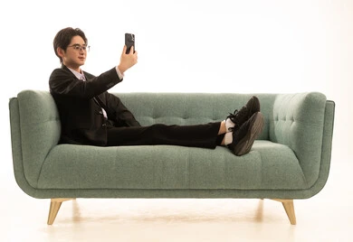 Taking a selfie with a mobile phone, gestures of happiness and joy, a portrait of a young Asian man wearing a formal suit sitting on the couch holding a phone in his hand, making a video call, white background.