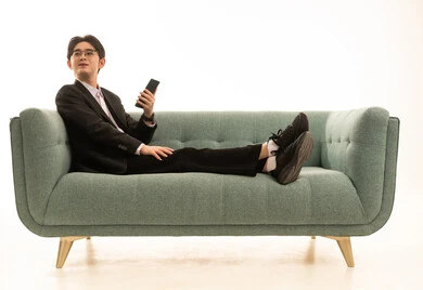 النظرالى شىء ما، استخدام جهاز تقني حديث و متطور لانجاز المشروع، بورتريه لشاب اسيوى يرتدى البدلة الرسمية يجلس على الاريكة يمسك بيده الهاتف المحمول، خلفية بيضاء