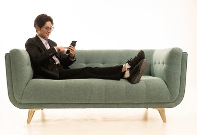 العمل عن بعد وتصفح الانترنت، استخدام جهاز تقني حديث و متطور لانجاز المشروع، بورتريه لشاب اسيوى يرتدى البدلة الرسمية يجلس على الاريكة يمسك بيده الهاتف المحمول، خلفية بيضاء