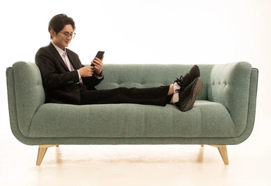 بورتريه لشاب اسيوى يرتدى البدلة الرسمية يجلس على الاريكة يمسك بيده الهاتف المحمول، العمل عن بعد وتصفح الانترنت، استخدام جهاز تقني حديث و متطور لانجاز المشروع،خلفية بيضاء