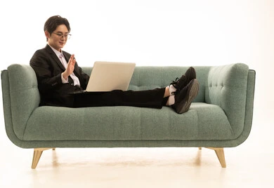 العمل عن بعد وتصفح الانترنت، استخدام جهاز تقني حديث و متطور لانجاز المشروع، بورتريه لشاب اسيوى يرتدى البدلة الرسمية يجلس على الاريكة يقوم باجراء مكالمة فيديو عبر الحاسوب، خلفية بيضاء