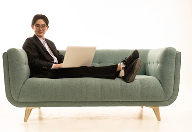 النظر الى الكاميرا بتعابير السرور، استخدام جهاز تقني حديث و متطور لانجاز المشروع، العمل عن بعد، بورتريه لشاب اسيوى يرتدى البدلة الرسمية يجلس على الاريكة يستخدم جهاز الحاسوب المحمول، خلفية بيضاء