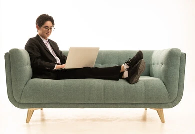 استخدام جهاز تقني حديث و متطور لانجاز المشروع، العمل عن بعد، بورتريه لشاب اسيوى يرتدى البدلة الرسمية يجلس على الاريكة يستخدم جهاز الحاسوب المحمول بايماءات السرور، خلفية بيضاء