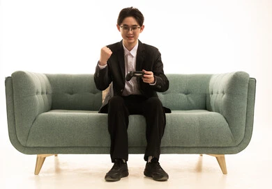 قضاء وقت ممتع في المنزل، استخدام التكنولوجيا الحديثة في الترفيه بورتريه لشاب اسيوى يرتدى البدلة الرسمية يجلس على كرسى مريح بعصا التحكم يرفع يده بايماءات الفوز والنصر، المتعة والتسلية والتفاعل ، خلفية بيضاء