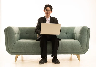 دمج التكنولوجيا بالعمل، إنجاز المهام المطلوبة عبر اللابتوب، بورتريه لشاب اسيوى يرتدى البدلة الرسمية يجلس على الاريكة يتمسك بيده الحاسوب الحمول، خلفية بيضاء