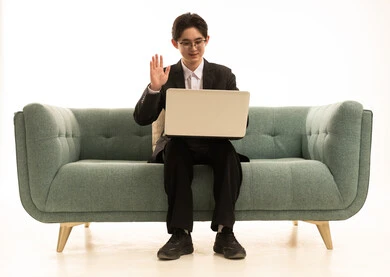 Greeting and peace, conducting online interviews, using modern technology to browse images, a portrait of an Asian young man wearing a formal suit sitting on the couch making a video call via laptop, white background.