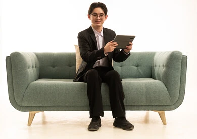 Looking at the camera with expressions of joy, conducting online interviews, using modern technology to browse images, a portrait of an Asian young man wearing a formal suit sitting on a sofa holding a tablet in his hand, white background.