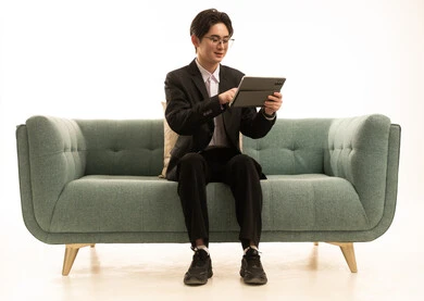 الدردشة مع الاقارب و الاصدقاء, القيام بمقابلات أونلاين، استخدام جهاز تقني حديث لتصفح الصور، بورتريه لشاب اسيوى يرتدى البدلة الرسمية يجلس على الاريكة يستخدم التابلت، خلفية بيضاء