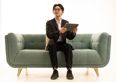 النظر الى شىء ما, القيام بمقابلات أونلاين، استخدام جهاز تقني حديث لتصفح الصور، بورتريه لشاب اسيوى يرتدى البدلة الرسمية يجلس على الاريكة يستخدم التابلت، خلفية بيضاء