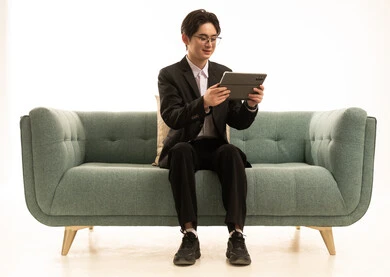 ايماءات الاندماج و التركيز، التواصل و الدردشة مع الاقارب, القيام بمقابلات أونلاين، استخدام جهاز تقني حديث لتصفح الصور، بورتريه لشاب اسيوى يرتدى البدلة الرسمية يجلس على الاريكة يستخدم التابلت، خلفية بيضاء