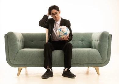 بورتريه لشاب اسيوى يرتدى البدلة الرسمية يحمل كورة الفريق يجلس على الاريكة ينظر الى الكاميرا و يضع يده على رأسه بنعابير الحسرة والحزن، متابعة مباراة كرة القدم، خلفية بيضاء