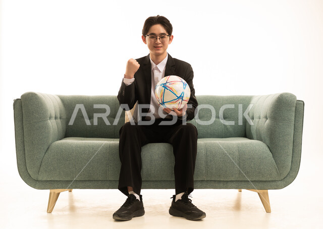 رفع قبضة اليد للاعلى بتعابير الفوز و الانتصار، بورتريه لشاب اسيوى يرتدى البدلة الرسمية يحمل كورة الفريق يجلس على الاريكة،  إيماءات الشغف والتشجيع للفريق، تشجيع فريق كرة قدم، خلفية بيضاء