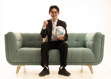 رفع قبضة اليد للاعلى بتعابير الفوز و الانتصار، بورتريه لشاب اسيوى يرتدى البدلة الرسمية يحمل كورة الفريق يجلس على الاريكة،  إيماءات الشغف والتشجيع للفريق، تشجيع فريق كرة قدم، خلفية بيضاء