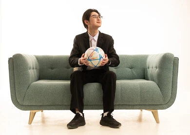 النظر الى شىء ما، بورتريه لشاب اسيوى يرتدى البدلة الرسمية يحمل كورة الفريق يجلس على الاريكة،  إيماءات الشغف والتشجيع للفريق، تشجيع فريق كرة قدم، خلفية بيضاء