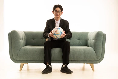 بورتريه لشاب اسيوى يرتدى البدلة الرسمية يحمل كورة الفريق يجلس على الاريكة،  إيماءات الشغف والتشجيع للفريق، تشجيع فريق كرة قدم، خلفية بيضاء