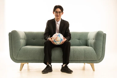 تشجيع فريق كرة قدم، بورتريه لشاب اسيوى يرتدى البدلة الرسمية يحمل كورة الفريق يجلس على الاريكة،  إيماءات الشغف والتشجيع للفريق، خلفية بيضاء